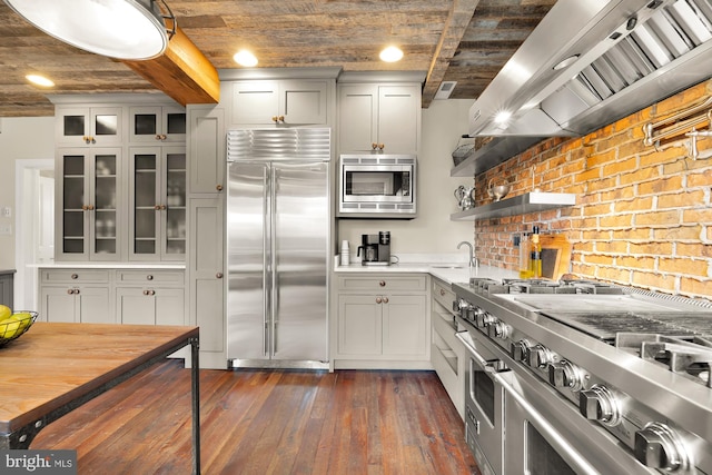 kitchen with glass insert cabinets, dark wood-type flooring, built in appliances, light countertops, and wall chimney range hood