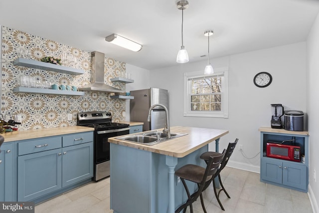 kitchen with an island with sink, butcher block counters, wall chimney exhaust hood, stainless steel appliances, and open shelves