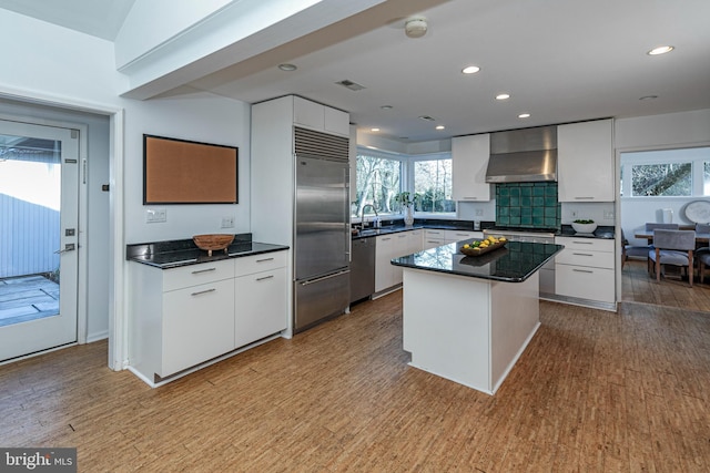 kitchen featuring appliances with stainless steel finishes, light wood-type flooring, white cabinetry, a kitchen island, and wall chimney exhaust hood