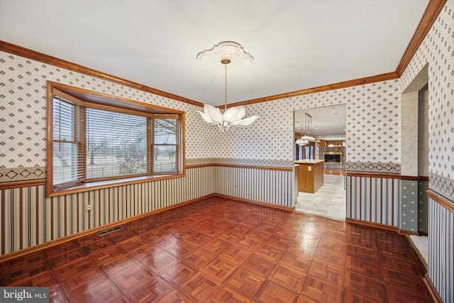 empty room featuring a wainscoted wall, visible vents, ornamental molding, a glass covered fireplace, and wallpapered walls