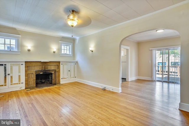 unfurnished living room featuring baseboards, arched walkways, ornamental molding, light wood-type flooring, and a brick fireplace