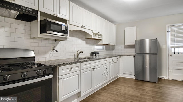 kitchen with dark stone countertops, stainless steel appliances, under cabinet range hood, white cabinetry, and a sink