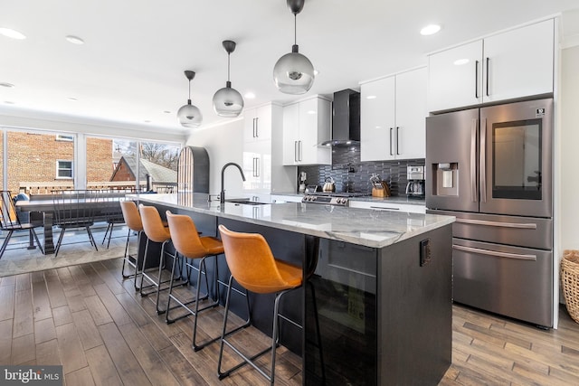 kitchen with dark wood-style floors, a center island with sink, stainless steel refrigerator with ice dispenser, wall chimney exhaust hood, and backsplash