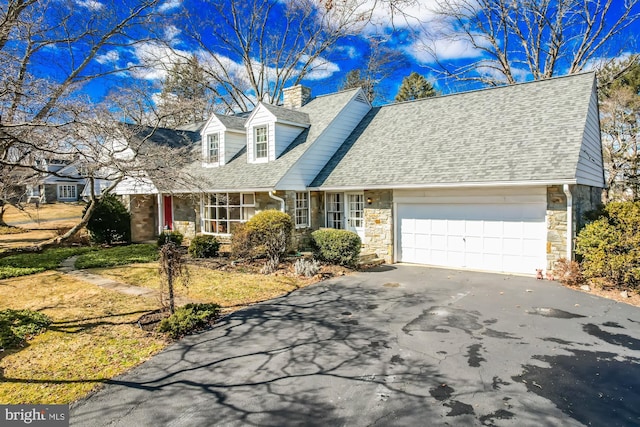 new england style home with an attached garage, a shingled roof, driveway, stone siding, and a chimney