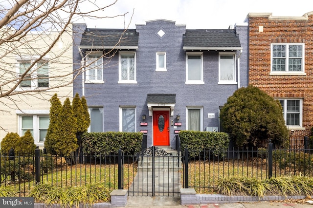 multi unit property with a fenced front yard, roof with shingles, brick siding, and mansard roof