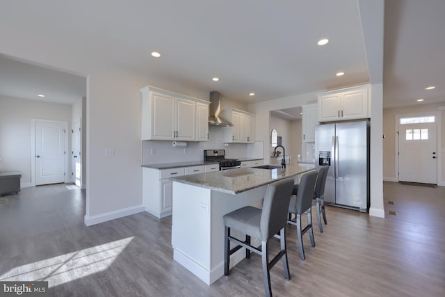 kitchen featuring a breakfast bar, a kitchen island with sink, appliances with stainless steel finishes, wall chimney range hood, and white cabinets