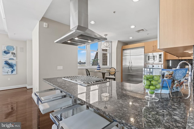 kitchen featuring island range hood, dark stone countertops, built in appliances, light brown cabinets, and pendant lighting