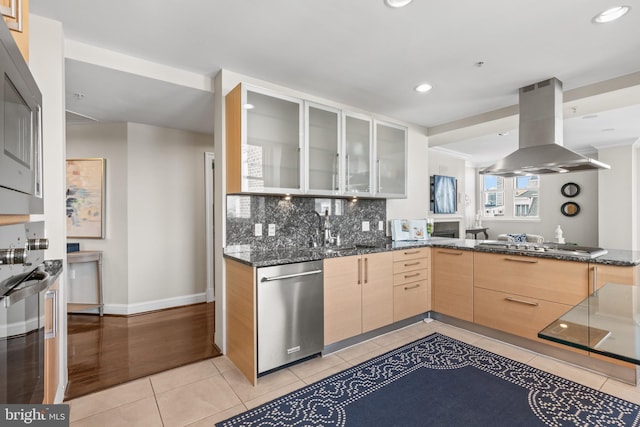 kitchen with dark stone counters, glass insert cabinets, appliances with stainless steel finishes, island exhaust hood, and light brown cabinets