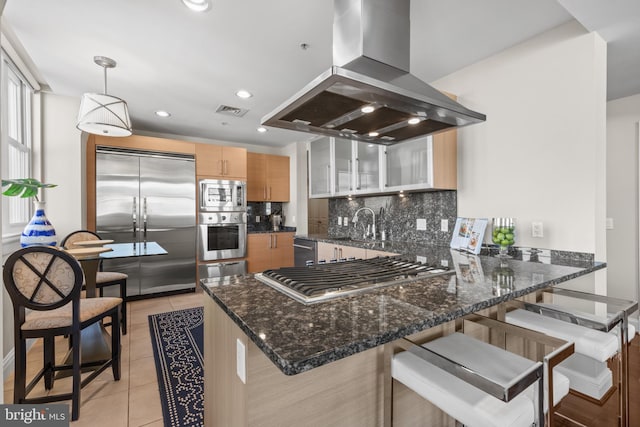 kitchen featuring island exhaust hood, glass insert cabinets, built in appliances, a peninsula, and a kitchen breakfast bar