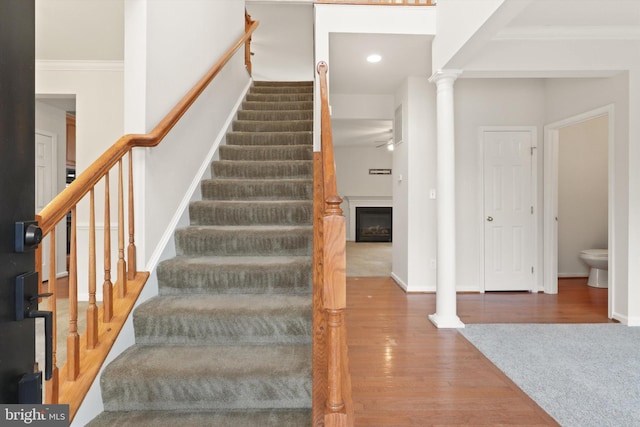 staircase featuring crown molding, a fireplace, decorative columns, a ceiling fan, and wood finished floors