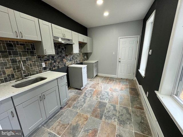 kitchen with baseboards, a sink, tasteful backsplash, stone tile flooring, and under cabinet range hood