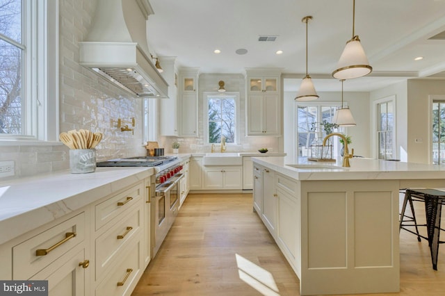 kitchen with custom range hood, glass insert cabinets, hanging light fixtures, a kitchen island with sink, and a sink