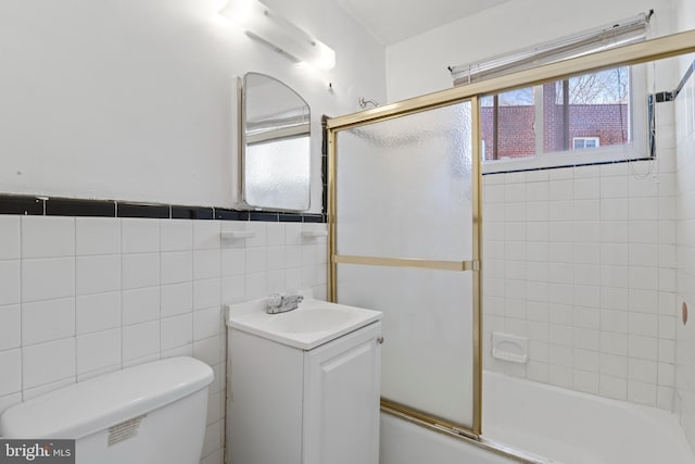 full bath featuring toilet, a wainscoted wall, vanity, tile walls, and combined bath / shower with glass door