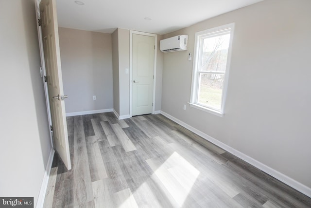 unfurnished bedroom featuring multiple windows, a wall mounted air conditioner, light wood-style flooring, and baseboards