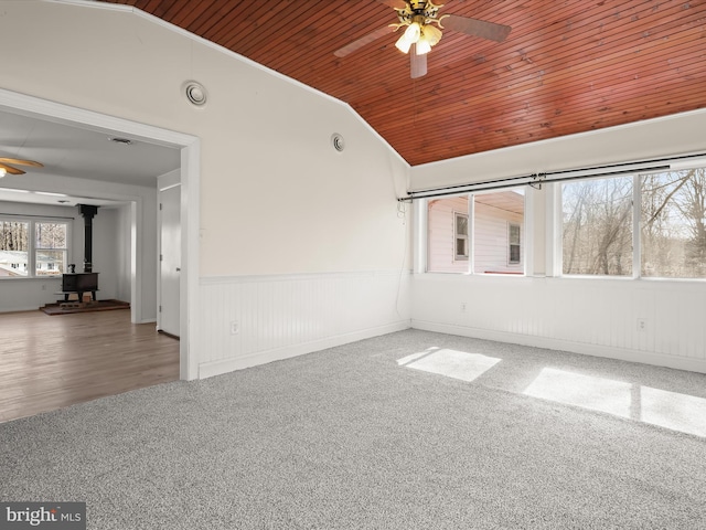 carpeted empty room featuring a ceiling fan, a wainscoted wall, lofted ceiling, a wood stove, and wooden ceiling