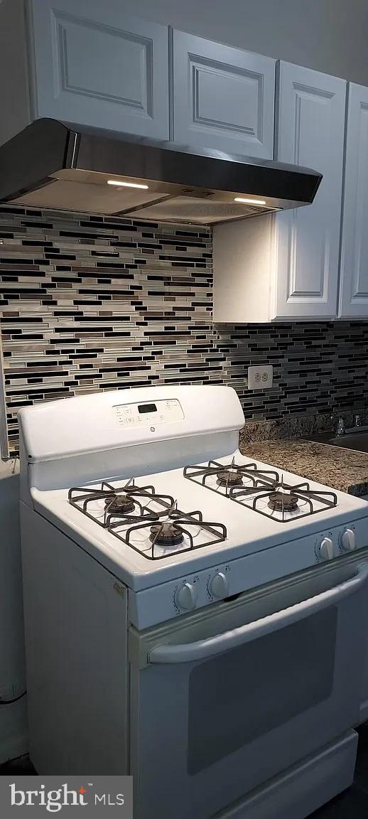 kitchen with white gas range, dark countertops, decorative backsplash, white cabinets, and exhaust hood