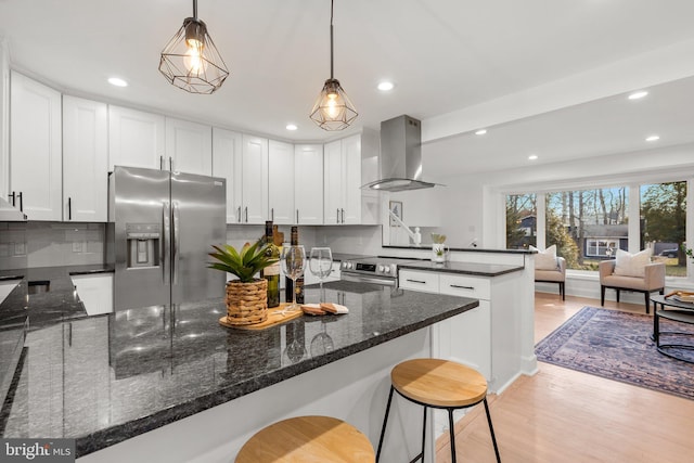 kitchen with stainless steel appliances, white cabinets, open floor plan, hanging light fixtures, and wall chimney range hood