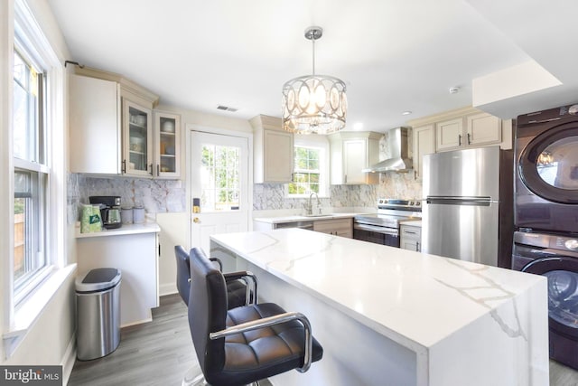 kitchen with wall chimney exhaust hood, appliances with stainless steel finishes, stacked washer / drying machine, a center island, and pendant lighting