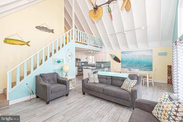 living room featuring beam ceiling, light wood finished floors, high vaulted ceiling, baseboards, and stairs