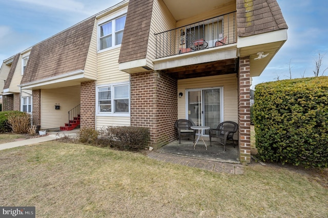 exterior space with mansard roof, a balcony, brick siding, a shingled roof, and a lawn