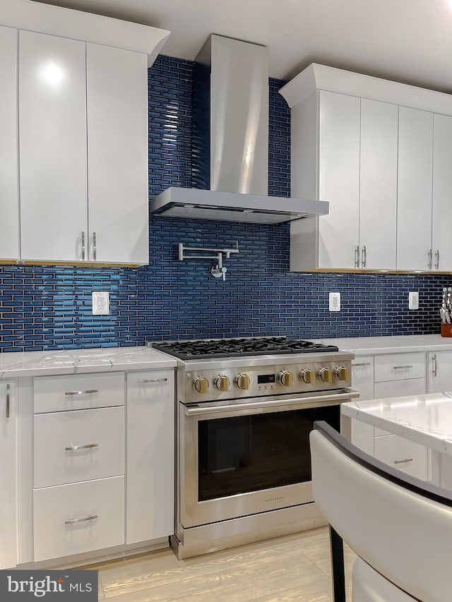 kitchen featuring wall chimney range hood, stainless steel gas range, backsplash, and white cabinetry