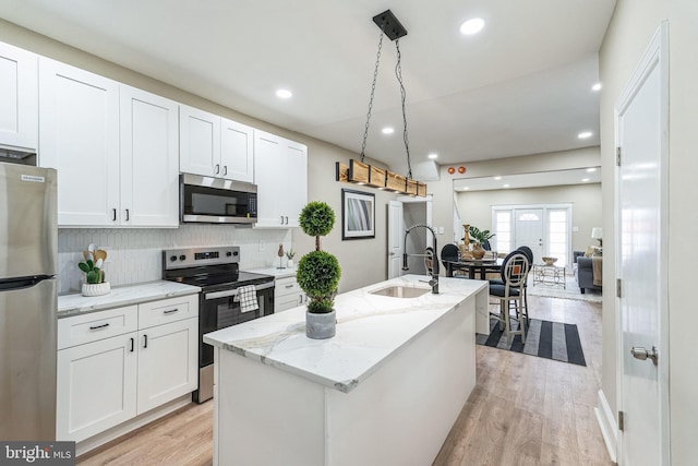 kitchen with light stone counters, decorative light fixtures, a center island with sink, stainless steel appliances, and white cabinetry