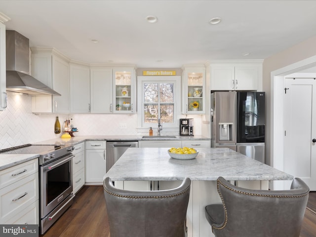 kitchen with stainless steel appliances, glass insert cabinets, a sink, wall chimney range hood, and a kitchen bar