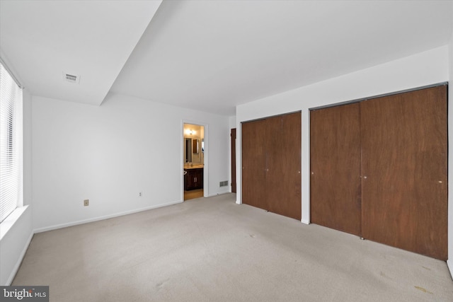 unfurnished bedroom featuring ensuite bathroom, light colored carpet, visible vents, baseboards, and multiple closets