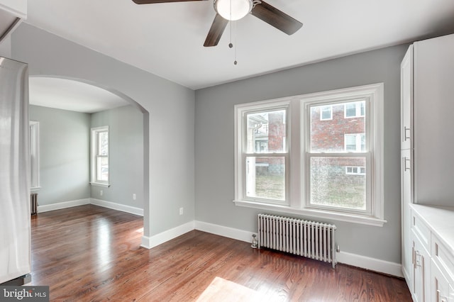 unfurnished room featuring arched walkways, radiator, a healthy amount of sunlight, and wood finished floors