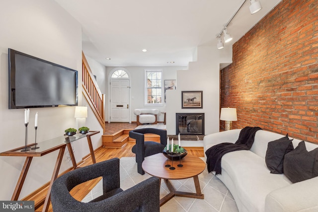 living room with brick wall, stairs, light wood-style flooring, rail lighting, and a glass covered fireplace