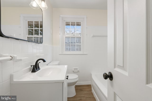 bathroom with a tub to relax in, toilet, wood finished floors, vanity, and tile walls