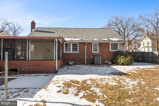 snow covered property with a sunroom, a chimney, fence, central AC, and brick siding