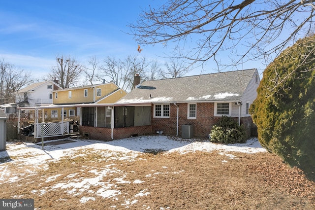 snow covered property with brick siding, roof with shingles, a chimney, a sunroom, and central AC