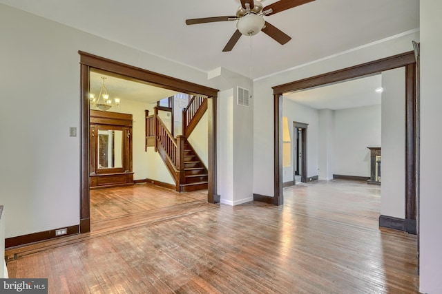 empty room featuring ceiling fan with notable chandelier, wood finished floors, visible vents, baseboards, and stairs