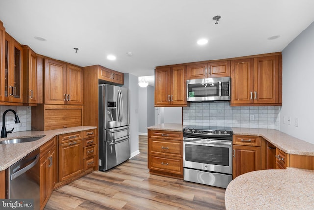 kitchen with stainless steel appliances, a sink, brown cabinets, light stone countertops, and glass insert cabinets