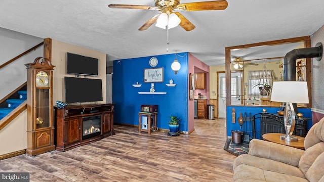 living area featuring wood finished floors, a ceiling fan, stairway, a glass covered fireplace, and a wood stove