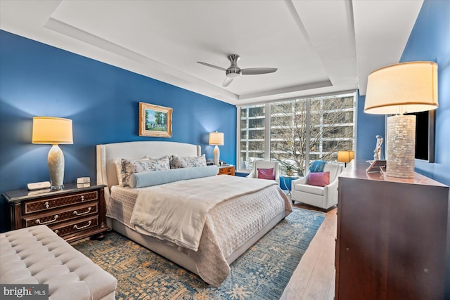 bedroom with ceiling fan, wood-type flooring, expansive windows, and a raised ceiling