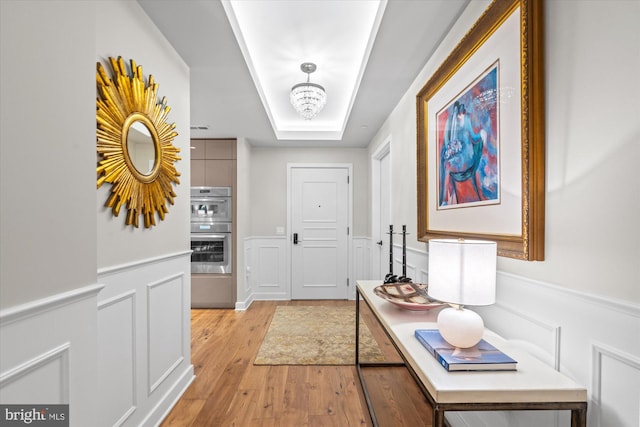 corridor featuring light wood finished floors, a raised ceiling, wainscoting, a decorative wall, and a notable chandelier