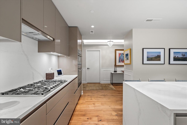 kitchen with under cabinet range hood, stainless steel gas cooktop, a wainscoted wall, light wood-style floors, and gray cabinets