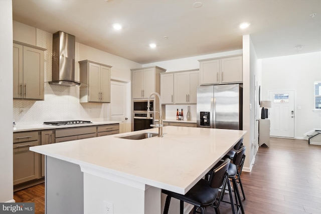 kitchen featuring gray cabinetry, an island with sink, a sink, stainless steel appliances, and wall chimney range hood