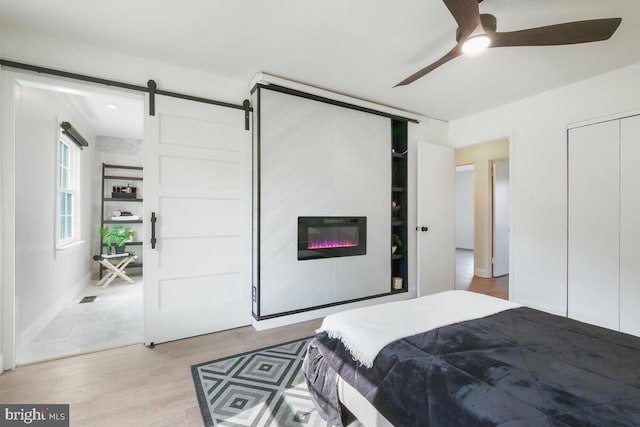 bedroom with a large fireplace, a barn door, light wood-type flooring, and a ceiling fan