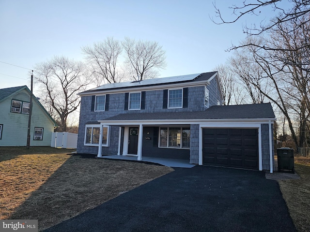 traditional-style house featuring roof with shingles, covered porch, an attached garage, roof mounted solar panels, and driveway