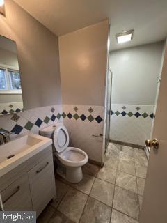 bathroom with a wainscoted wall, toilet, a stall shower, tile walls, and vanity