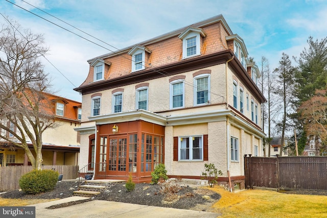 second empire-style home with a sunroom, fence, mansard roof, and stucco siding