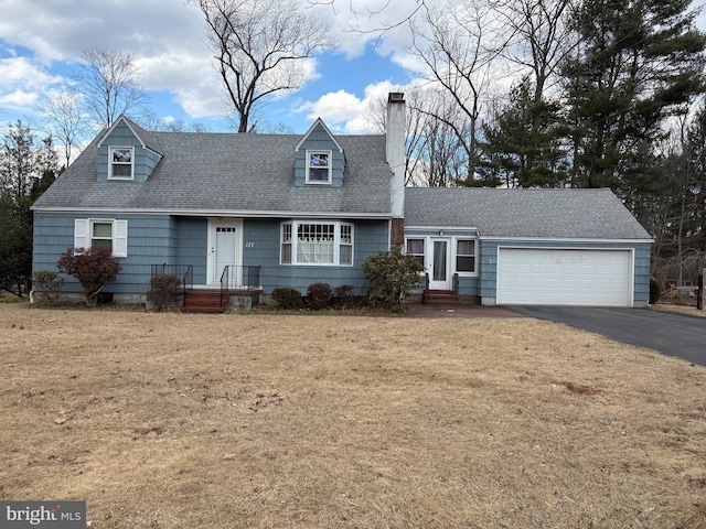 cape cod home with an attached garage, driveway, roof with shingles, a front lawn, and a chimney