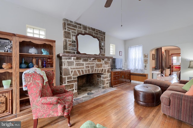 living area featuring arched walkways, a stone fireplace, wood finished floors, radiator heating unit, and plenty of natural light