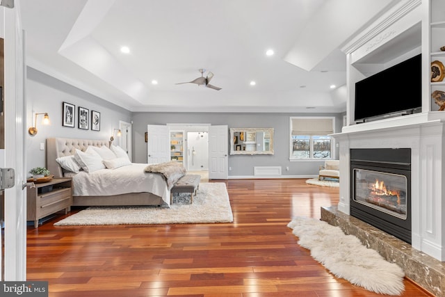 bedroom with a tray ceiling, recessed lighting, a glass covered fireplace, baseboards, and hardwood / wood-style flooring