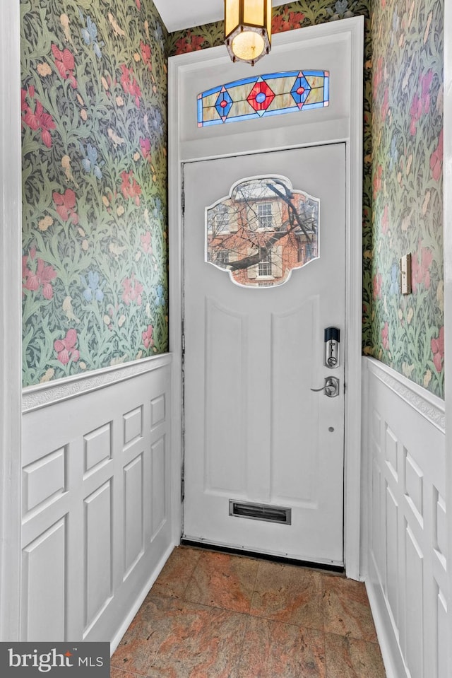 doorway to outside with wallpapered walls, stone finish floor, and wainscoting