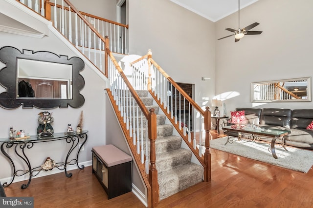 stairs featuring ceiling fan, a high ceiling, wood finished floors, baseboards, and ornamental molding