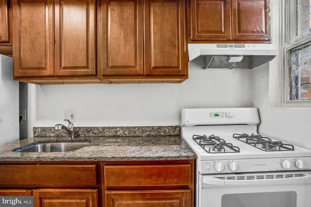 kitchen featuring stone countertops, under cabinet range hood, a sink, brown cabinetry, and gas range gas stove